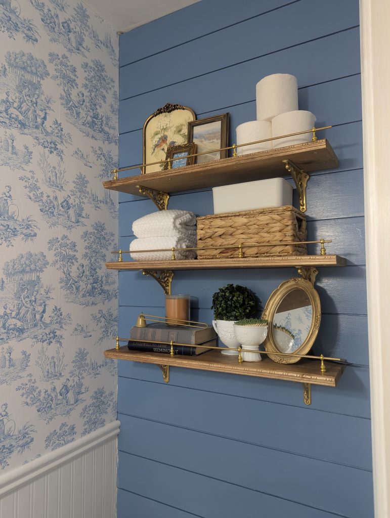 After: A fresh blue and white cottage powder room with beadboard "paneling", vintage style toile wallpaper, a much needed new toilet, gold mirror, blue painted shiplap accent wall, and upgraded shelving with ornate brackets.