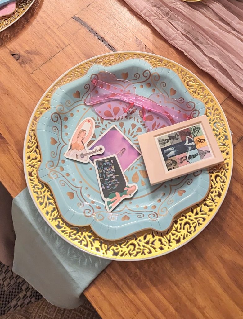 taylor swift table birthday party favors, gold rimmed plate with blue disposable plate, topped with an Instax frame, Taylor swift stickers, and pink heart sunglasses.